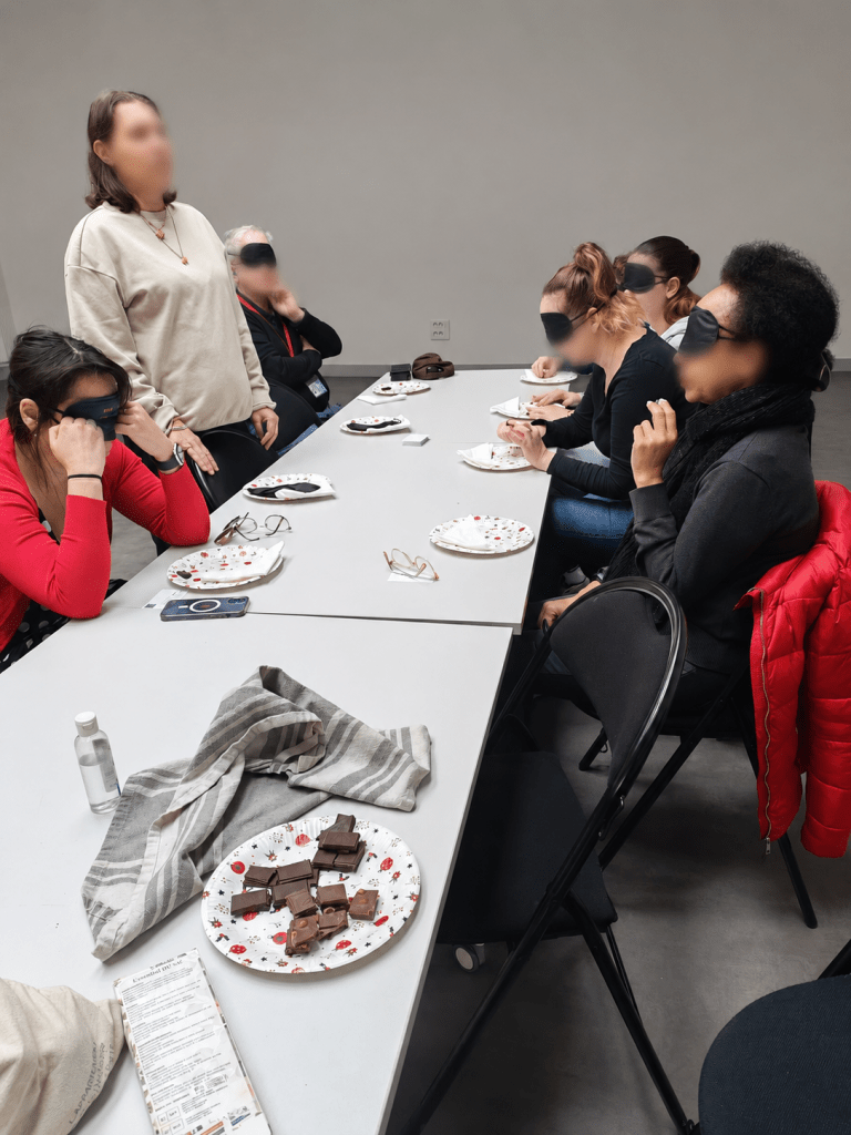 Un groupe de participantes prend part à un atelier sensoriel autour d’une longue table. Les yeux bandés, elles découvrent et dégustent différents morceaux de chocolat, guidées par une animatrice debout à leurs côtés. Des assiettes, des serviettes et des lunettes sont disposées sur la table. L’image transmet une ambiance conviviale, attentive et participative.