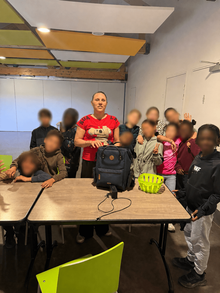 La photo montre un groupe d’enfants réunis autour d’une animatrice dans une grande salle d’activités. L’adulte, placée au centre, porte un tee-shirt rouge et se tient derrière une table sur laquelle sont posés un sac à dos bleu, un petit panier vert et un chargeur. Les enfants entourent l’animatrice dans une ambiance chaleureuse et conviviale. Certains se tiennent debout, d’autres s’appuient sur la table, et plusieurs font des gestes joyeux comme un signe de la main ou un signe de victoire. Leurs visages ont été floutés afin de préserver leur anonymat. La salle est lumineuse et moderne, avec de grands panneaux acoustiques colorés suspendus au plafond, dans des tons jaune, vert et blanc. L’ensemble donne l’image d’un moment collectif, dynamique et bienveillant, pris à la fin ou au cours d’un atelier avec un jeune public.